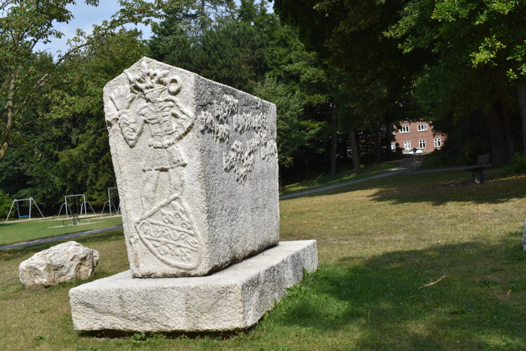 ’Bosnisk-Hercegovinsk-Dansk Stećak’ i Sankt Jørgens Park i Odense. I baggrunden kan Jomsborgvej skimtes mellem træerne. Den bosniske bjerglilje, der sjældent findes uden for Bosnien, pryder ’gavlen’, mens noget så odenseansk som et H.C. Andersen-citat pryder langsiden: ”Leve er ikke nok! Sagde han, ”Solskin, Frihed og en lille Blomst maa man have!”. På modsatte gavl hilser en skikkelse pænt på beskueren. Motivet er inspireret af Radimlja i Hercegovina. For skikkelsens fødder ligger et bosnisk skjold og et nedlagt spyd som symbol på fred. På den anden langside er der et citat fra digtet ’Modra rijeka – Blå flod’: ”… Over trætte, over bitre. Over tjørn, over tornbusk. Over hede og over hindring. Over forudanelse og over tvivl…” Mehmedalija ”Mak” Dizdar, der var fra Stolac, skrev digtet i 1960’erne og rammer den lidelse og tragedie, som de bosniske flygtninge har overlevet, men også det de fandt og fik i Danmark. (Jimmy Munk Larsen)